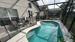 A covered swimming pool with a curved edge, surrounded by a fence and chairs, and a view of a house in the background. - Stay Awhile at the Fairways (Haines City)