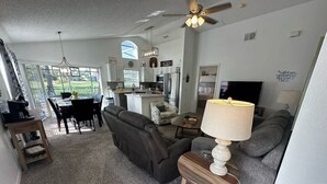 A spacious living room with a gray sofa, a wooden coffee table, and a ceiling fan. A kitchen is visible in the background with modern appliances. - Stay Awhile at the Fairways (Haines City)