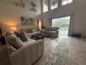 A spacious living room with a large sofa set, a coffee table, and a view of a swimming pool through large windows.