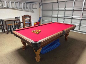 A pool table with balls, a blue cloth, and a wooden frame in a room with a garage door and chairs. - Golden Fairway Retreat (Haines City)