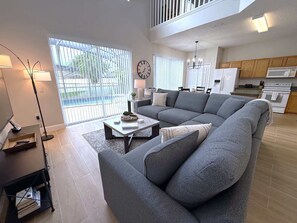 Ein modernes Wohnzimmer mit einem grauen Ecksofa, einem Couchtisch und einer Stehlampe. Es gibt einen Essbereich mit Tisch und Stühlen, sowie eine Küche mit Holzschränken und einem Kühlschrank.
