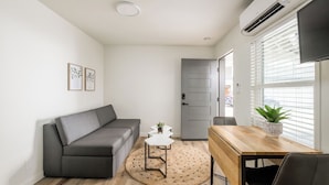 A modern living room with a grey sofa, a wooden dining table, a small white table, and a potted plant. - First Floor King Suite (101) (Oceanside)
