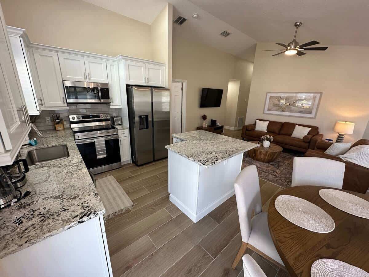 A modern kitchen with white cabinetry, stainless steel appliances, and a granite countertop. There is a dining area with a wooden table and chairs, and a living room with a brown sofa and a television.