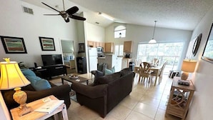A living room with a sofa, coffee table, and TV. There is a dining area with chairs and a table. The kitchen is visible in the background.