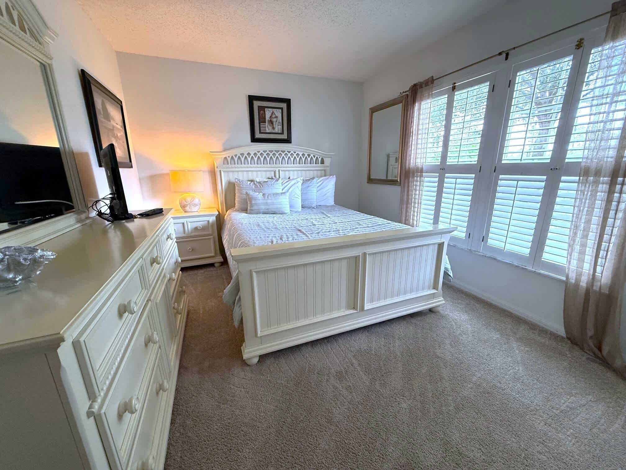 A bedroom with a large bed, a dresser, a nightstand, a lamp, a framed picture on the wall, and a window with curtains.