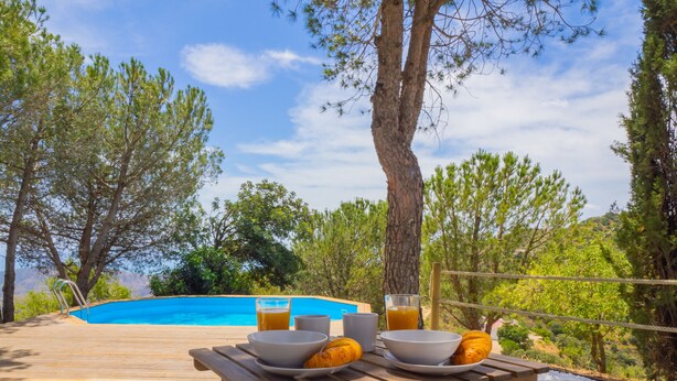 A wooden table with bowls, a glass of juice, and croissants on a pebble-covered ground near a pool.