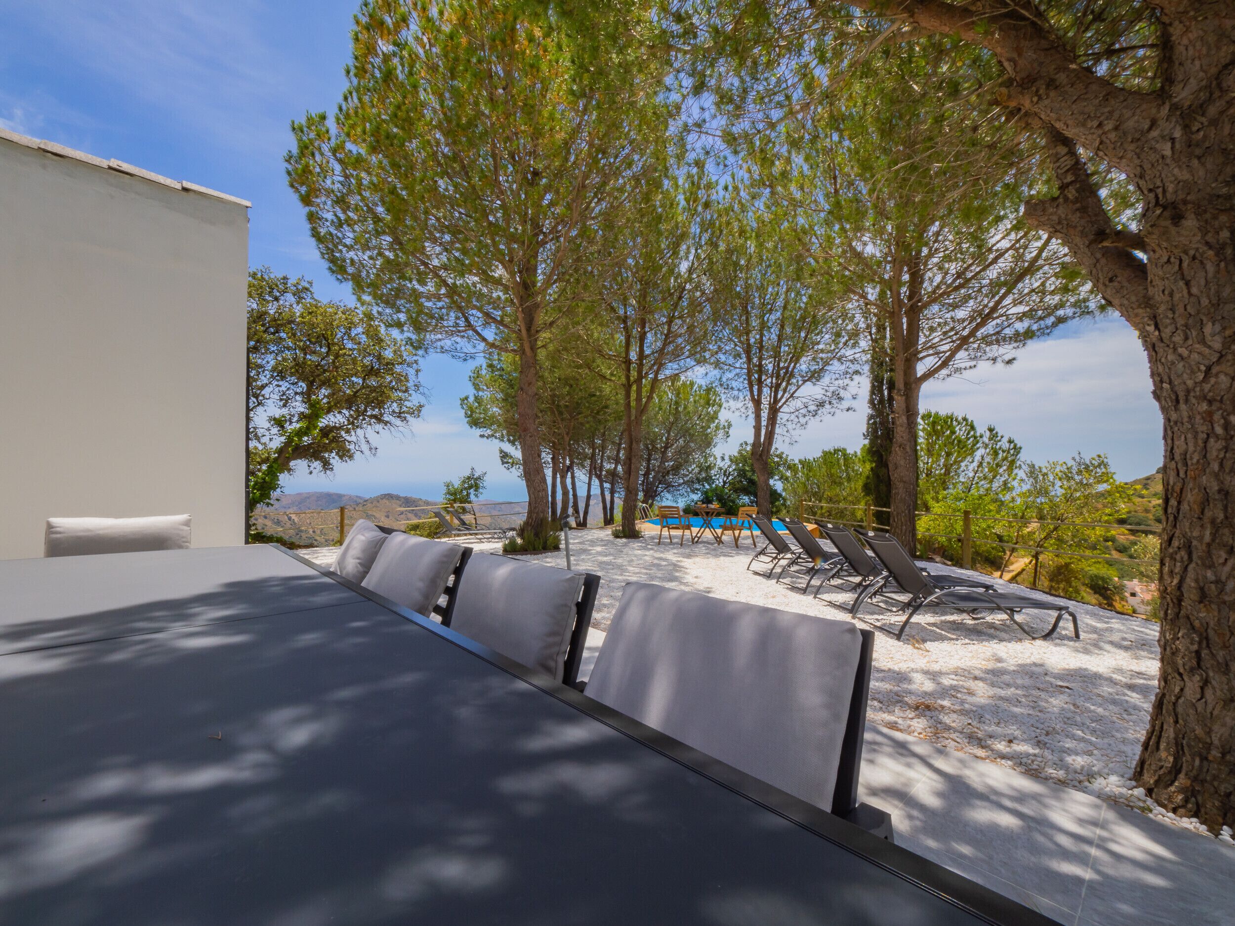 A sunlit outdoor seating area with a pool and lounge chairs, surrounded by trees and a view of the landscape.