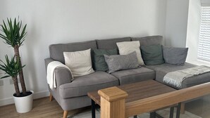 A modern living room with a grey sectional sofa, a wooden coffee table, a potted plant, and a large window with blinds. - Bay View House (Bangor)