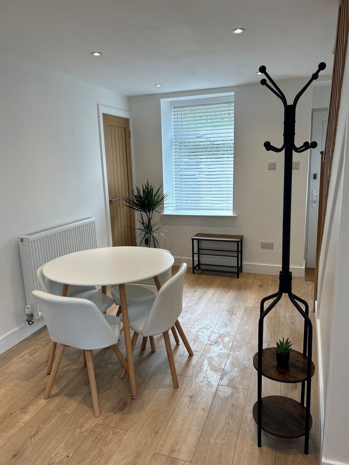 A modern dining area with a round table and white chairs, a radiator, a window with blinds, a bench, and a coat rack with a plant.