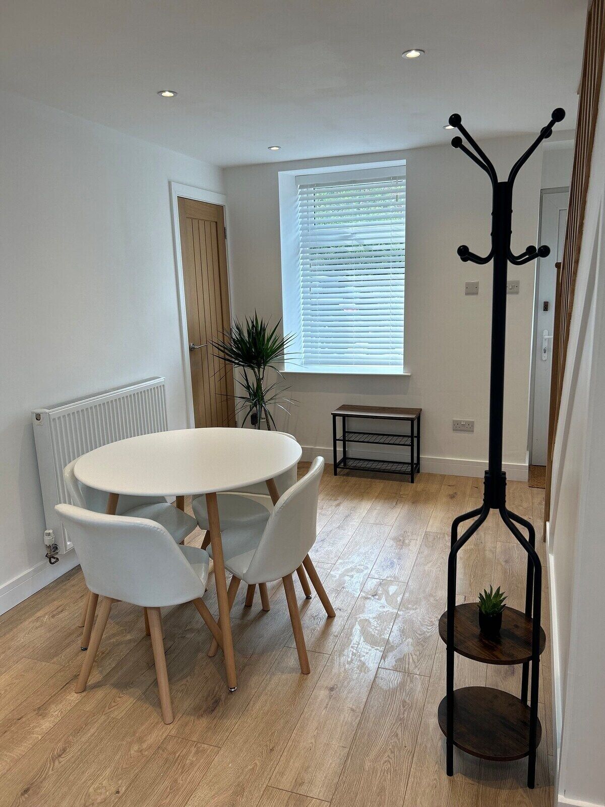A modern dining area with a round table and white chairs, a radiator, a window with blinds, a bench, and a coat rack with a plant.