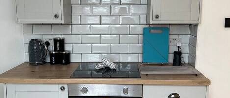 Una cocina compacta con gabinetes blancos, una campana extractora de acero inoxidable, un horno integrado y una tabla de cortar azul sobre la encimera.