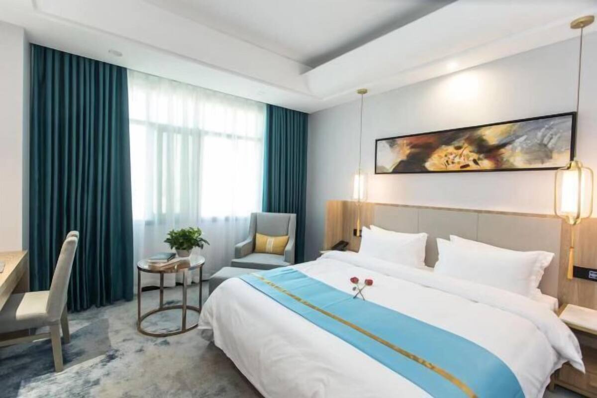A hotel room with a large bed, a desk, a chair, a small table with a plant, and a painting on the wall.
