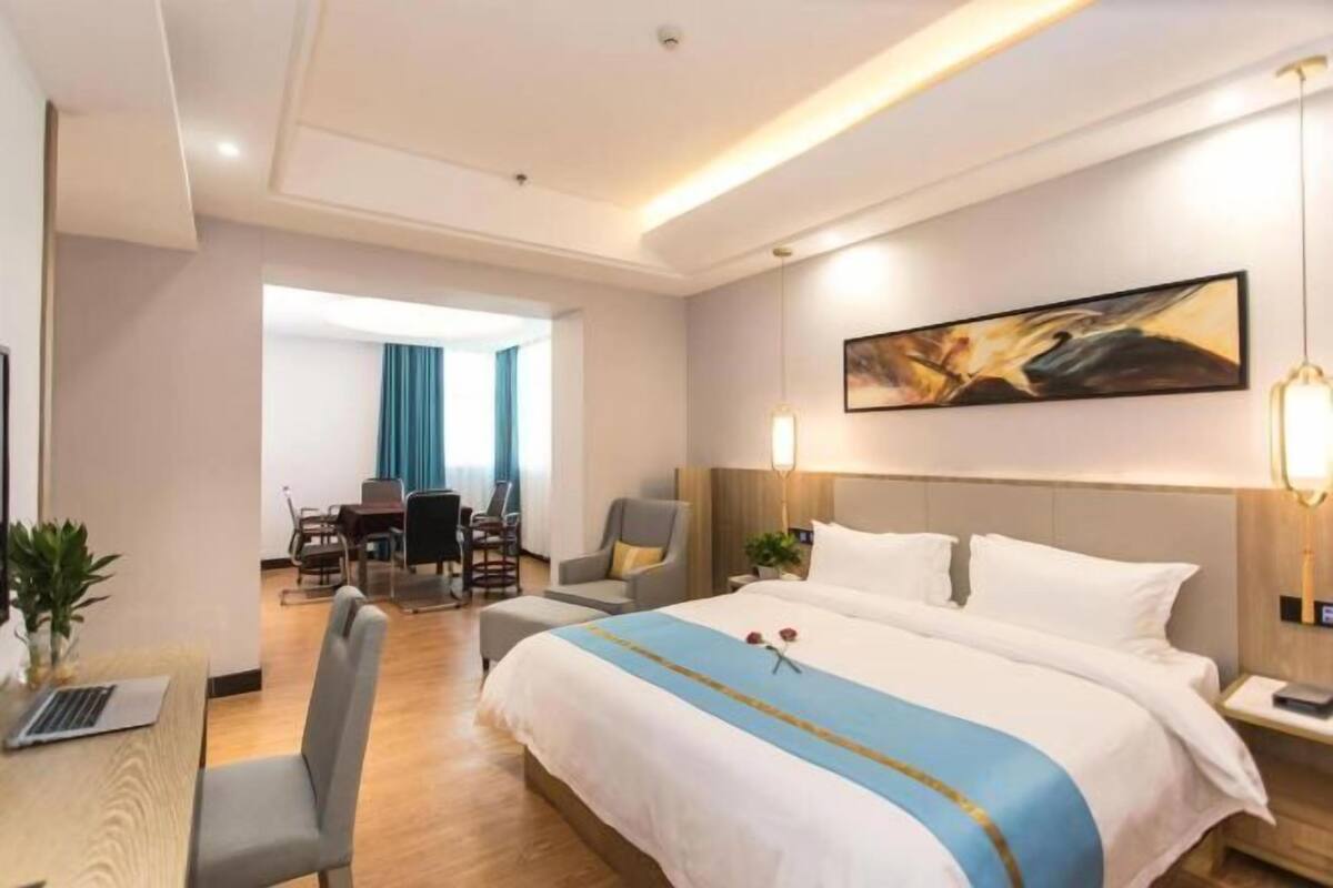 A hotel room with a large bed, a desk with a laptop, a dining area with chairs, and a painting on the wall.