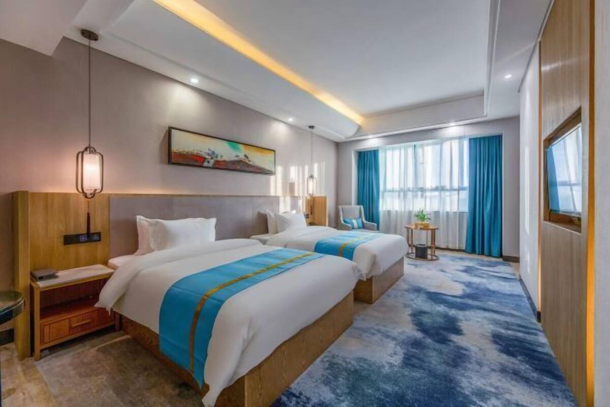 A hotel room with two beds, a desk, a chair, a small table with a plant, and a painting on the wall.