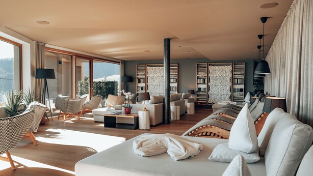 A spacious living room with a large sofa, a coffee table, and a bookshelf. There is a large window providing natural light and a view of the outdoors. - Panoramahotel Watles (Malles Venosta)