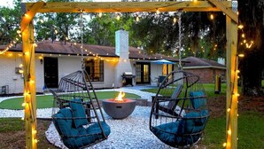 Un jardin avec un foyer, deux chaises suspendues et une pergola illuminée de guirlandes lumineuses.