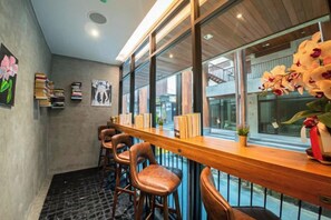 A bar area with wooden stools and a long counter, a potted plant, and a window with a view of the outside. - J4 Samui Hotel (Koh Samui)