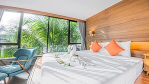 A hotel room with a large bed, a blue armchair, and a view of palm trees through large windows. - J4 Samui Hotel (Koh Samui)