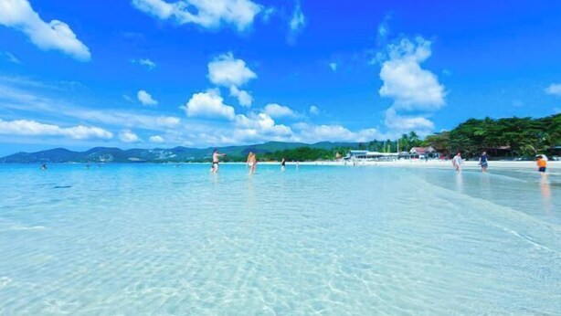 A beach with clear turquoise water, people swimming and walking on the shore, and a distant mountain range under a blue sky with clouds. - J4 Samui Hotel (Koh Samui)