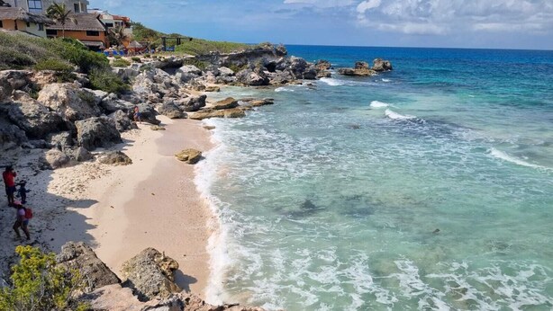 Una playa con aguas turquesas cristalinas, costa rocosa y algunas personas caminando por la orilla.