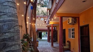 Un pasillo estrecho con luces colgantes, una palmera y un edificio de techo rojo con una puerta.