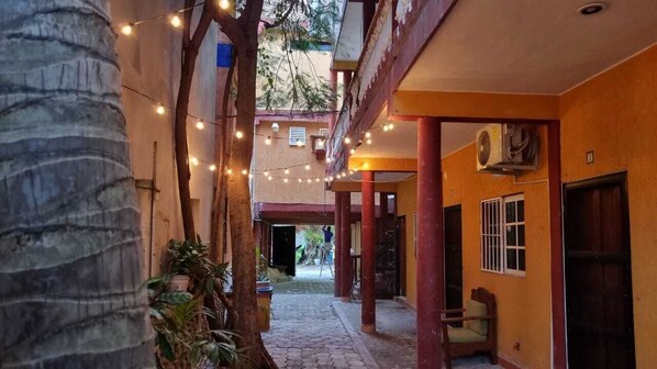 Un pasillo estrecho con luces colgantes, una palmera y un edificio de techo rojo con una puerta.