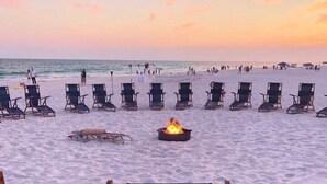 Een strand met rijen ligbedden en een vuurkorf in het midden, met het logo 'Surfside Resort' op de leuning van de stoelen.