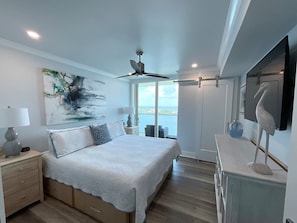 Une chambre à coucher avec un lit, des tables de chevet, un ventilateur de plafond, un tableau et une vue sur la mer.