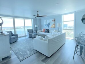 Un salon moderne avec un canapé blanc, une table basse et un coin repas avec des chaises. Une grande fenêtre offre une vue sur l’océan.