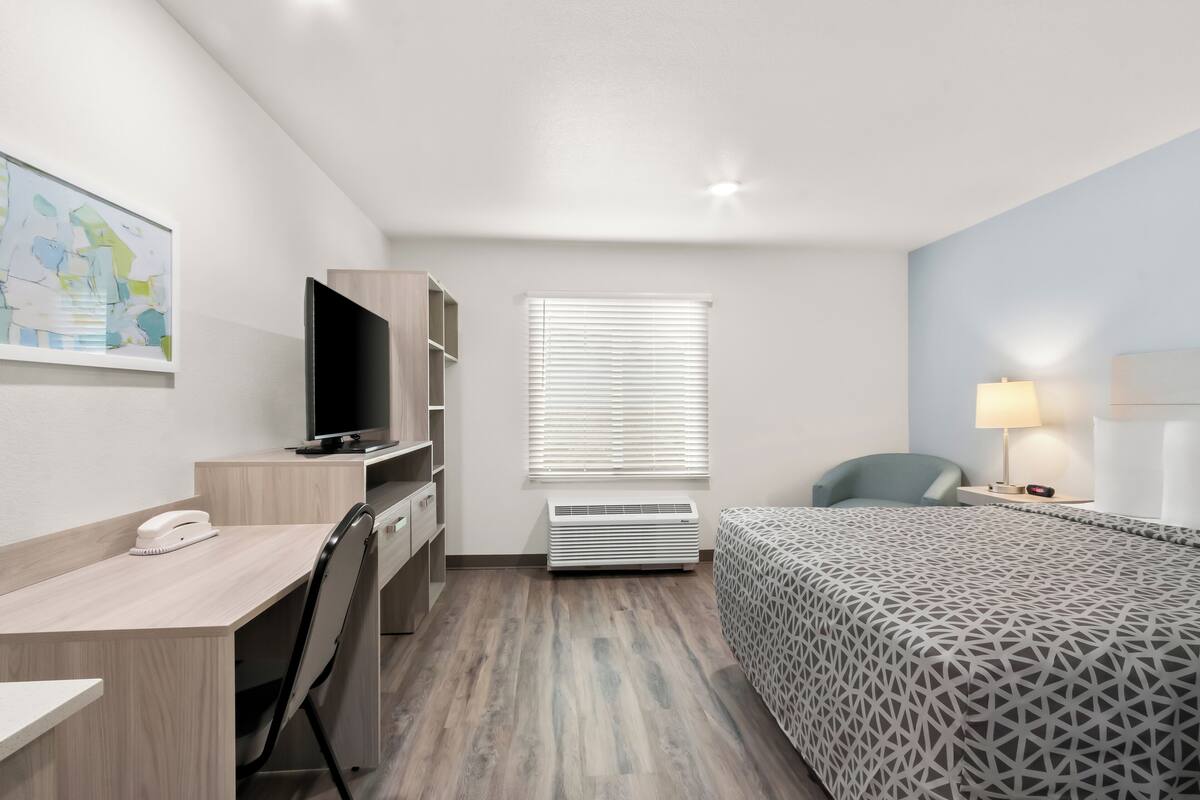 A hotel room with a desk, chair, bed, TV, air conditioning unit, and a window with blinds.
