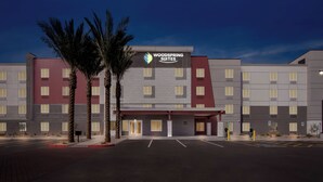 Un hotel de varios pisos con un cartel llamativo de Woodspring Suites, un estacionamiento y palmeras.