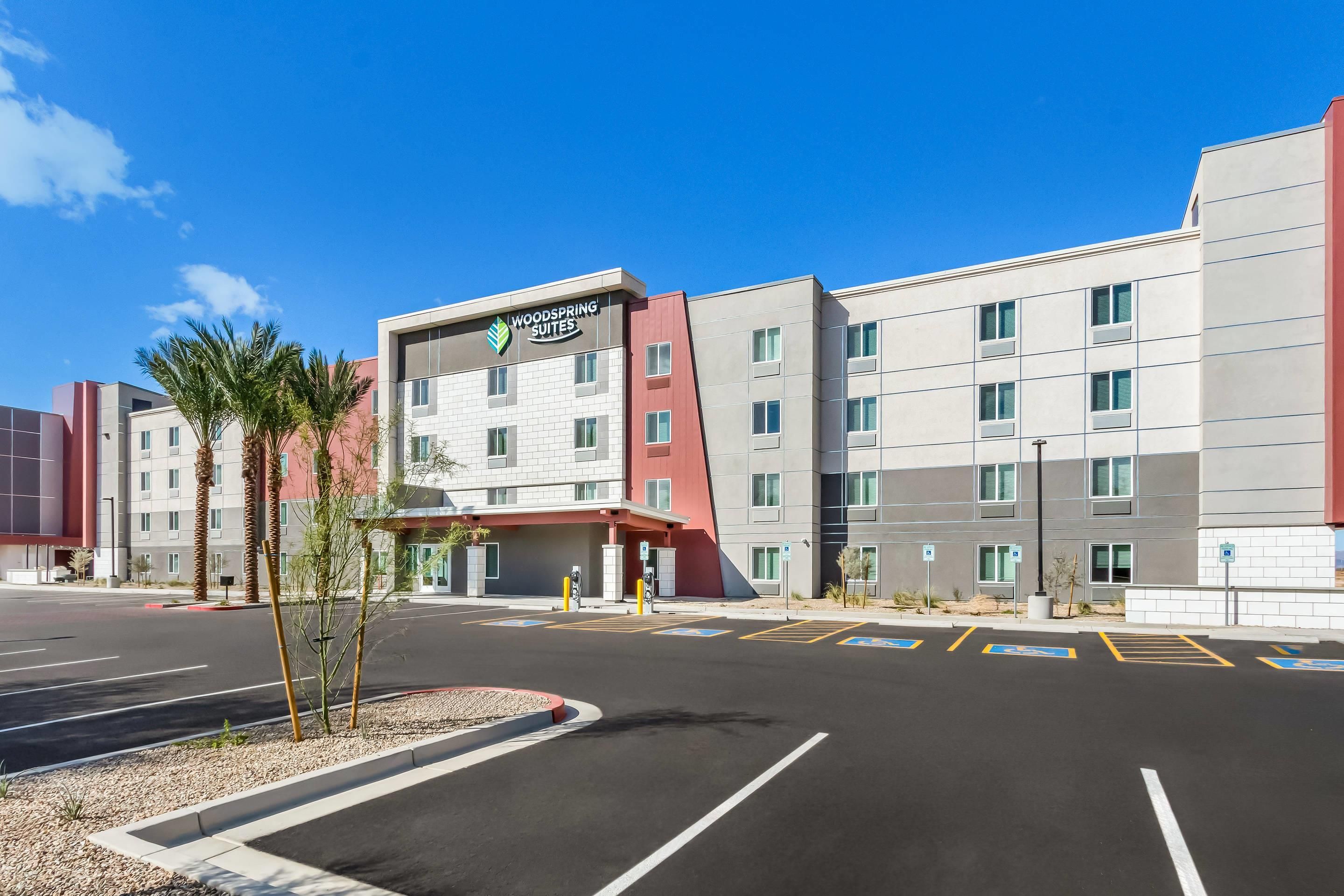 A multi-story hotel with a prominent 'Woodspring Suites' sign, parking spaces, and a clear blue sky.