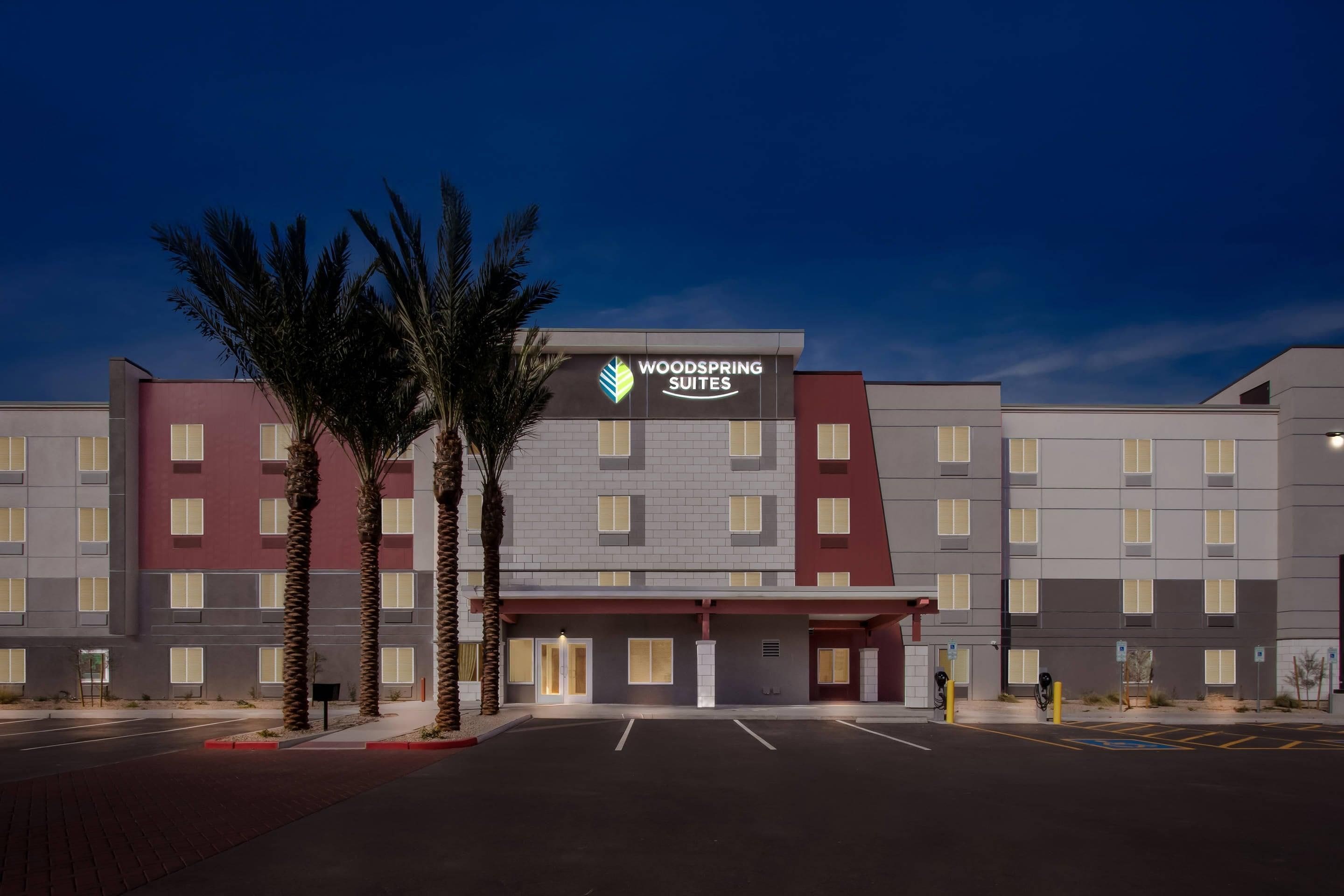 Un hotel con un llamativo letrero de Woodspring Suites, que presenta un diseño moderno con techo plano y una combinación de paredes exteriores en rojo y gris.