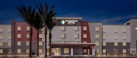 Un hotel con un llamativo letrero de Woodspring Suites, que presenta un diseño moderno con techo plano y una combinación de paredes exteriores en rojo y gris.
