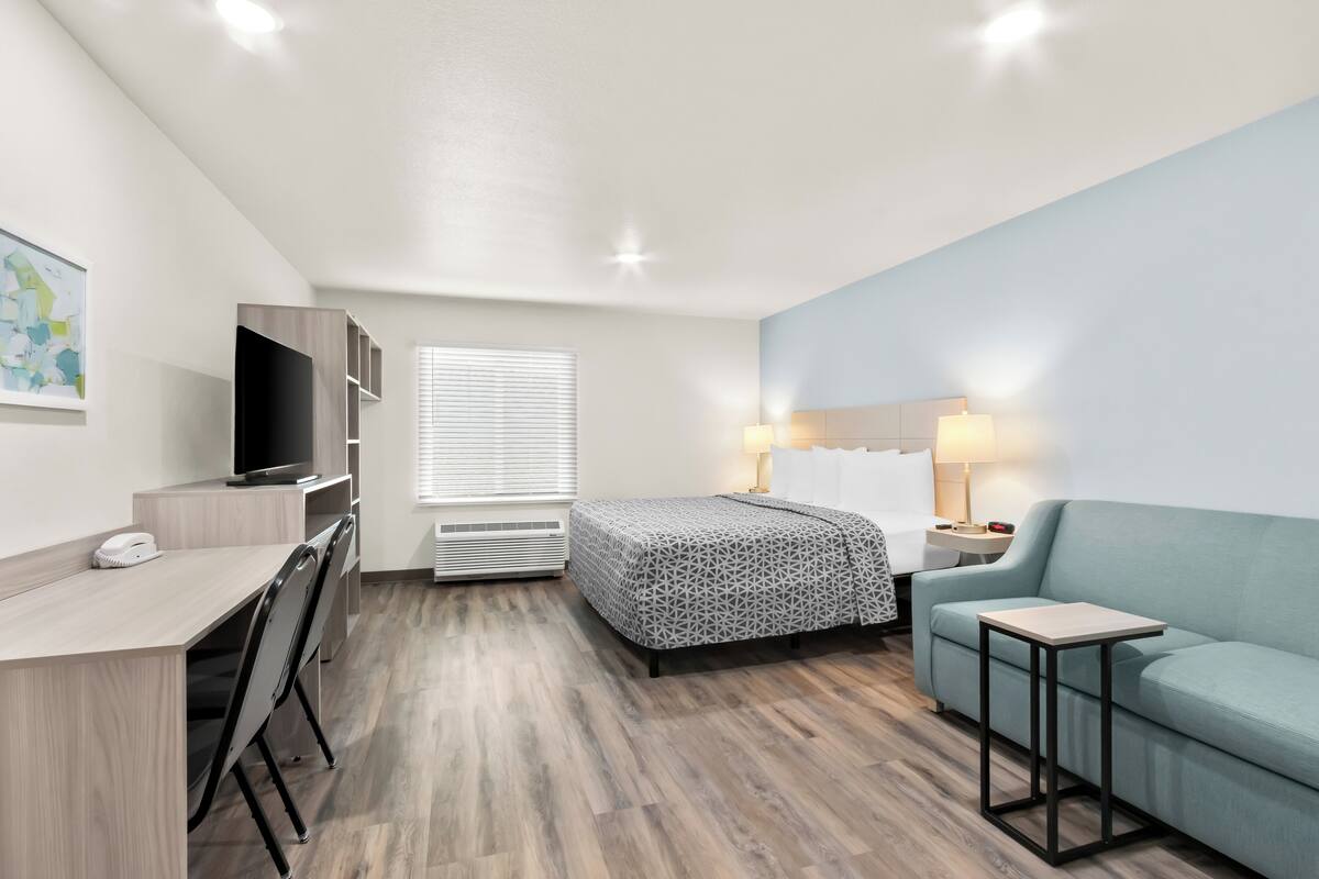 A modern hotel room with a bed, a desk, a TV, a sofa, and a small table.
