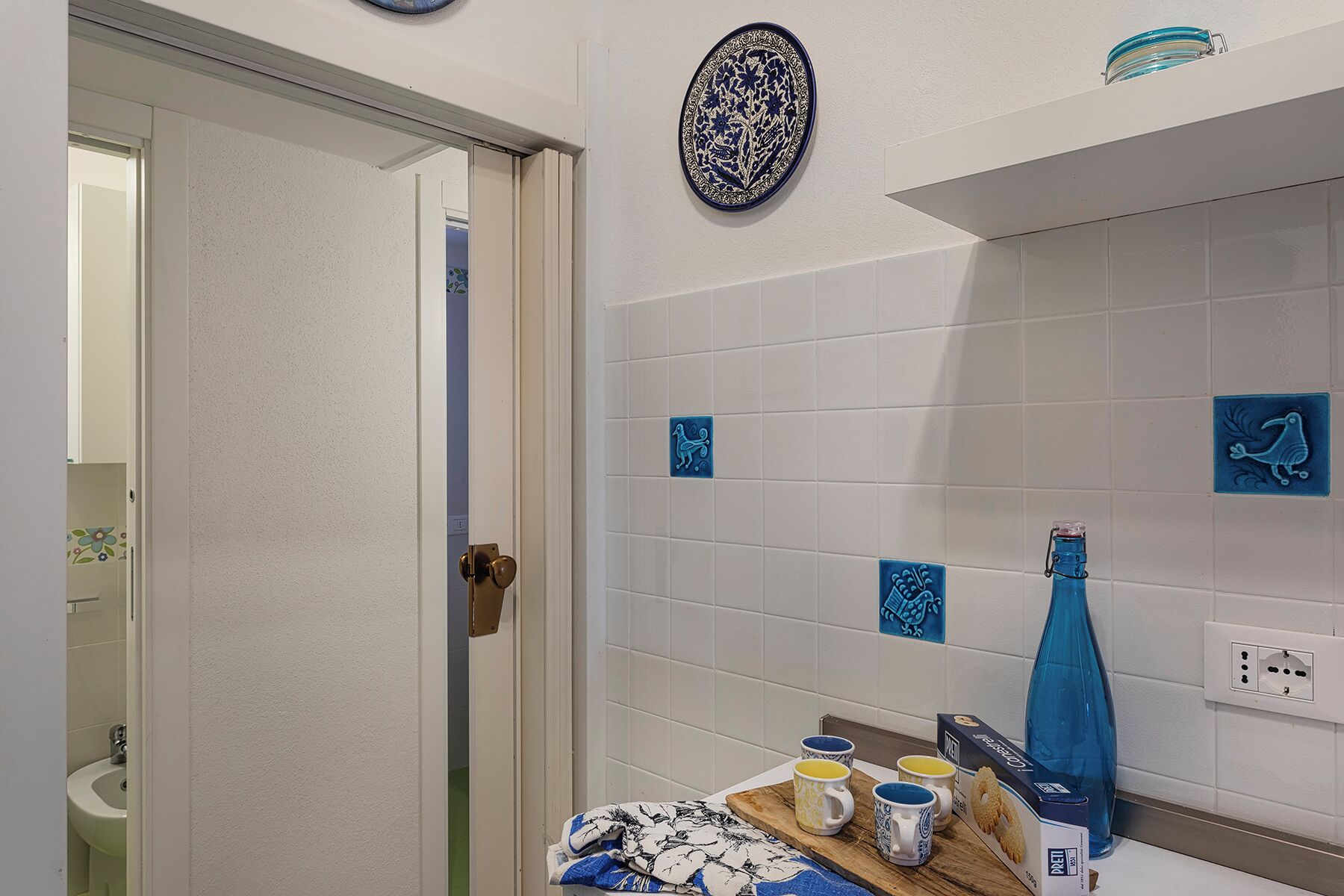 A bathroom with white tiles, a door, a mirror, and a small table with mugs and a box.
