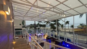A covered outdoor dining area with tables and chairs, a wooden deck, and a view of the beach and ocean. - Hotel Araguaia (Praia Grande)
