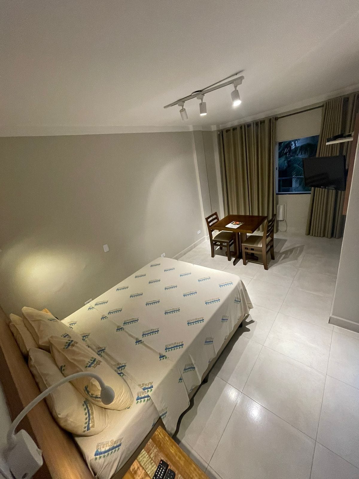 Habitación de hotel con cama, escritorio y silla. Se observa una ventana con cortinas y un televisor montado en la pared.