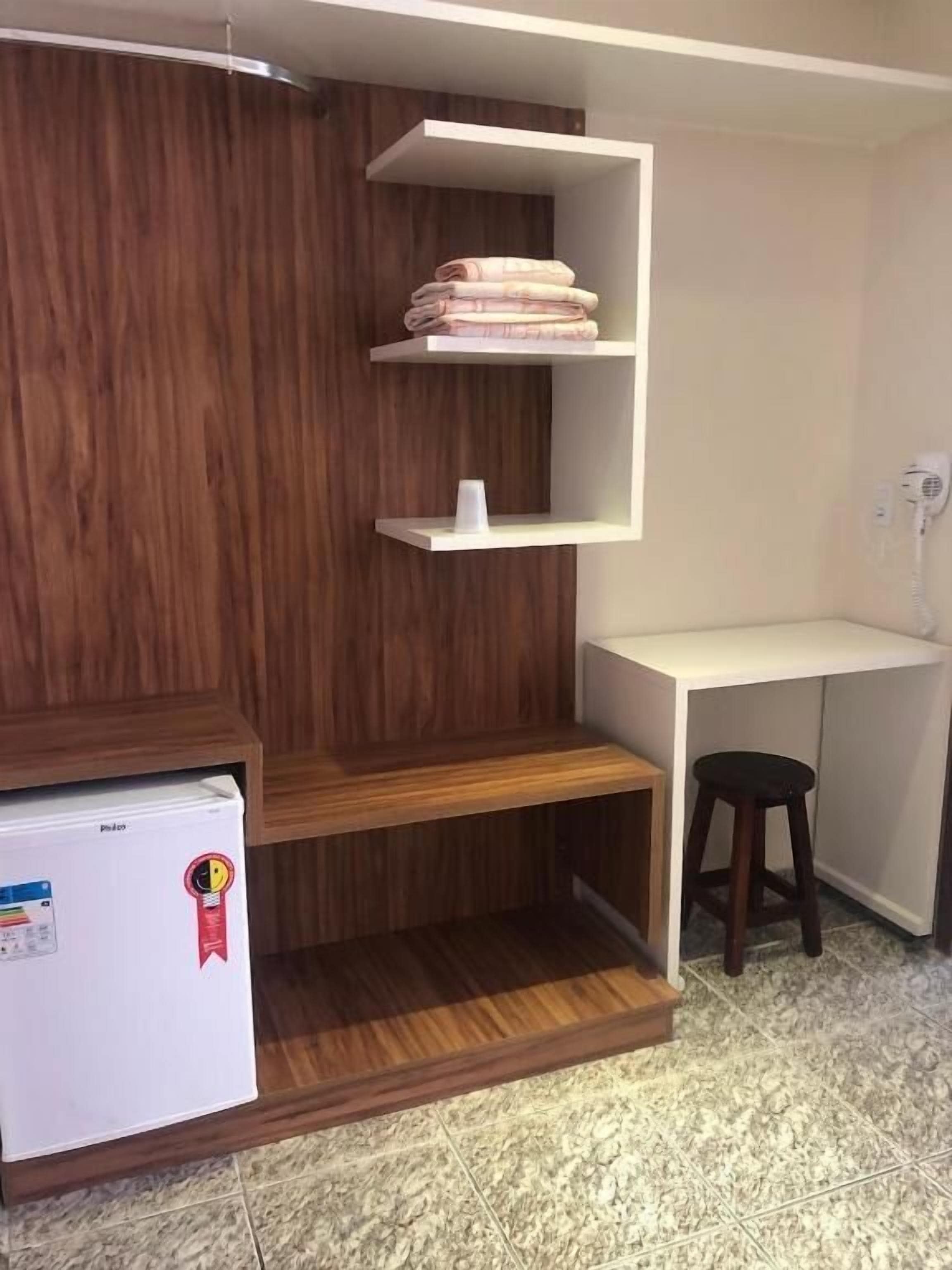 A compact room with a wooden wardrobe, a white desk, a stool, and a small refrigerator.