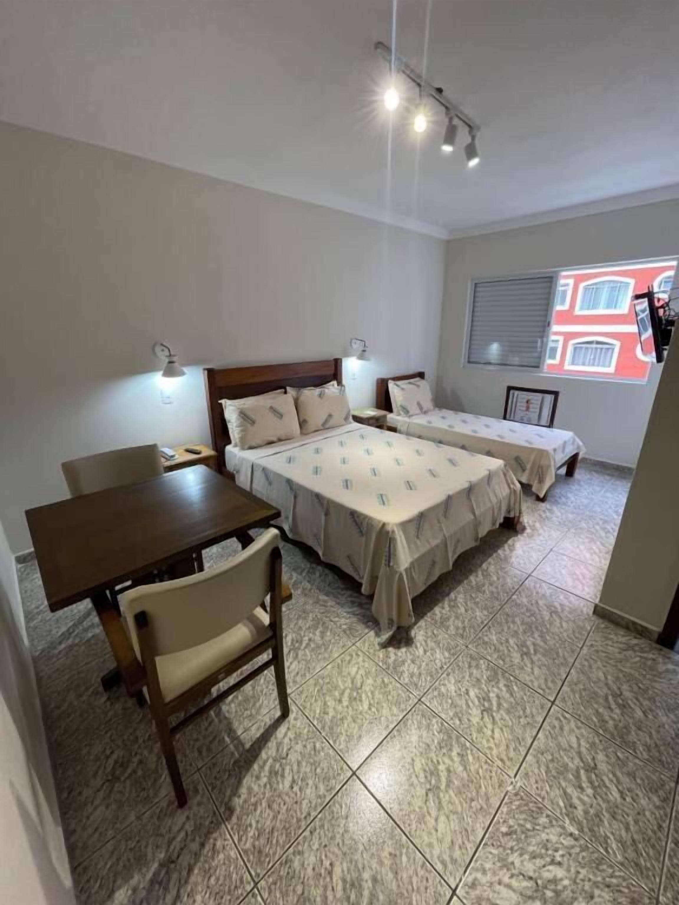 A room with two beds, a table, and chairs. There are wall-mounted lights and a window with a view of buildings.