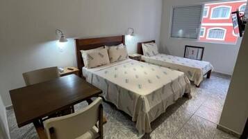 A room with two beds, a table, and chairs. There are wall-mounted lights and a window with a view of buildings.
