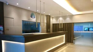 A modern reception desk with a logo, a glass partition, and pendant lighting. - Hotel Araguaia (Praia Grande)
