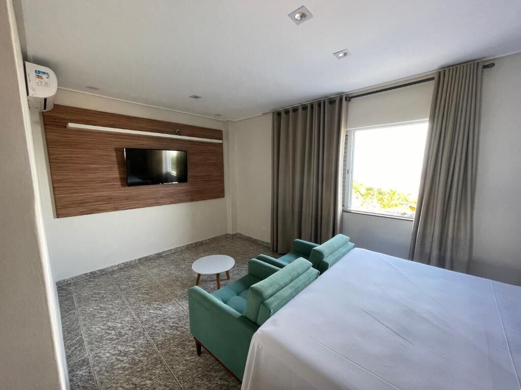 A hotel room with a bed, a green sofa, a small round table, a flat-screen TV mounted on a wooden panel, and a window with curtains.