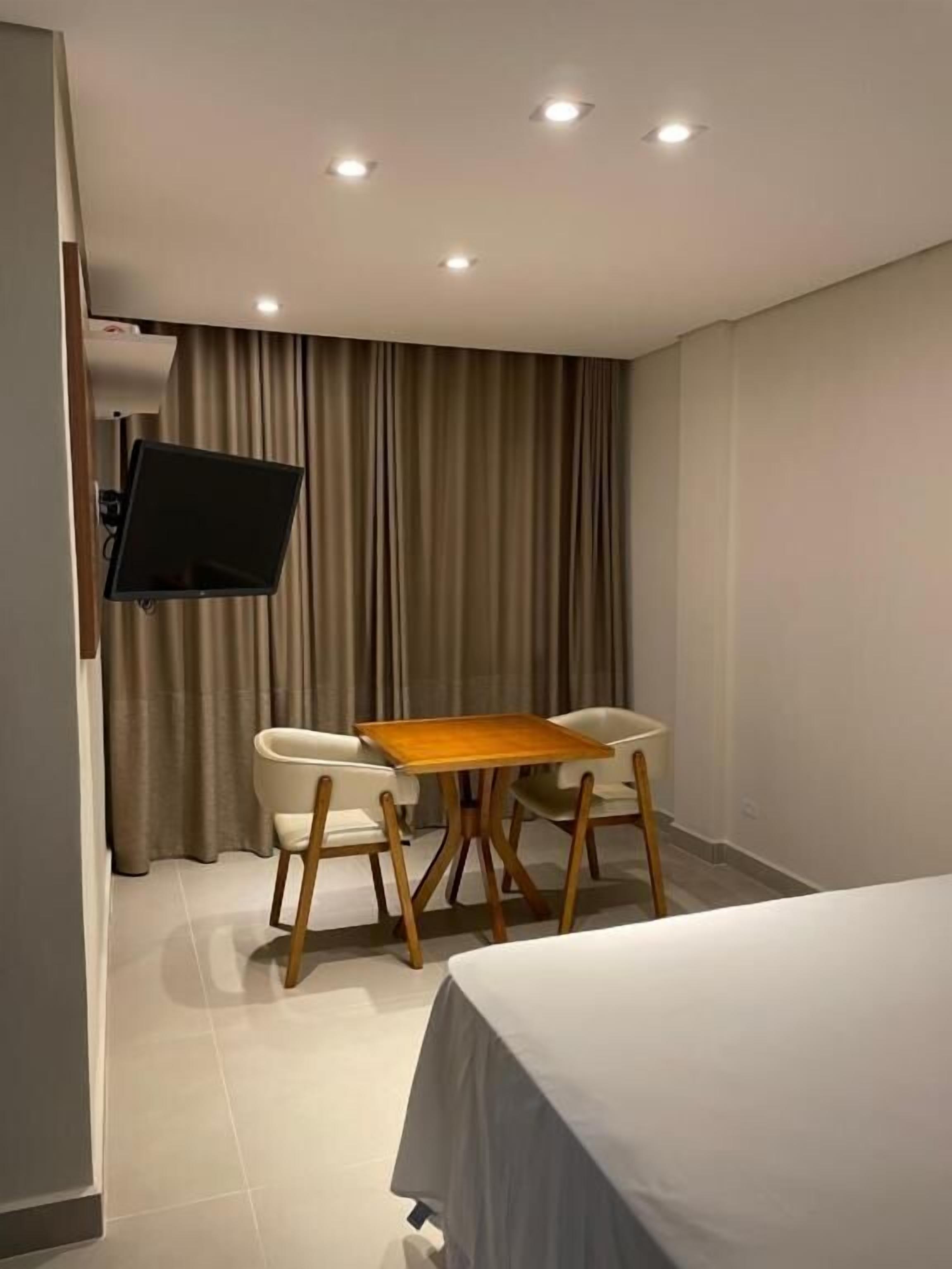 A modern hotel room with a flat-screen TV mounted on the wall, a wooden table with two chairs, a bed with a grey bedspread, and a large window with curtains.
