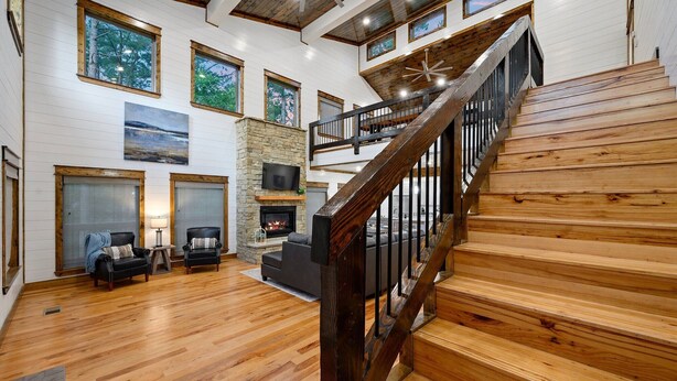 Un amplio salón con escalera de madera, chimenea de piedra y grandes ventanales con vistas al exterior.