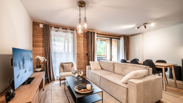 Ein modernes Wohnzimmer mit einer Couch, einem Couchtisch und einem Fernseher. Es gibt einen Essbereich mit Tisch und Stühlen.