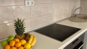Una cocina con mostrador blanco, un lavamanos y un frutero con manzanas, naranjas, plátanos y una piña.