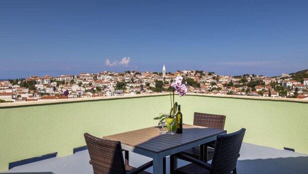Une table entourée de chaises en osier et une bouteille de vin, le tout sur un balcon donnant sur une ville côtière.
