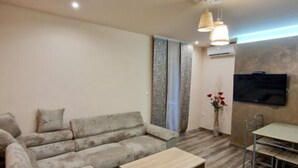 Una sala moderna con un sofá gris, una mesa de centro de madera, un televisor montado en la pared y una pequeña mesa de comedor con sillas.