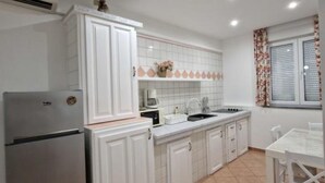 A compact kitchen with white cabinets, a built-in oven, and a window with floral curtains. - Pansion Belvedere - One Bedroom (Magnolia) (Mali Losinj)